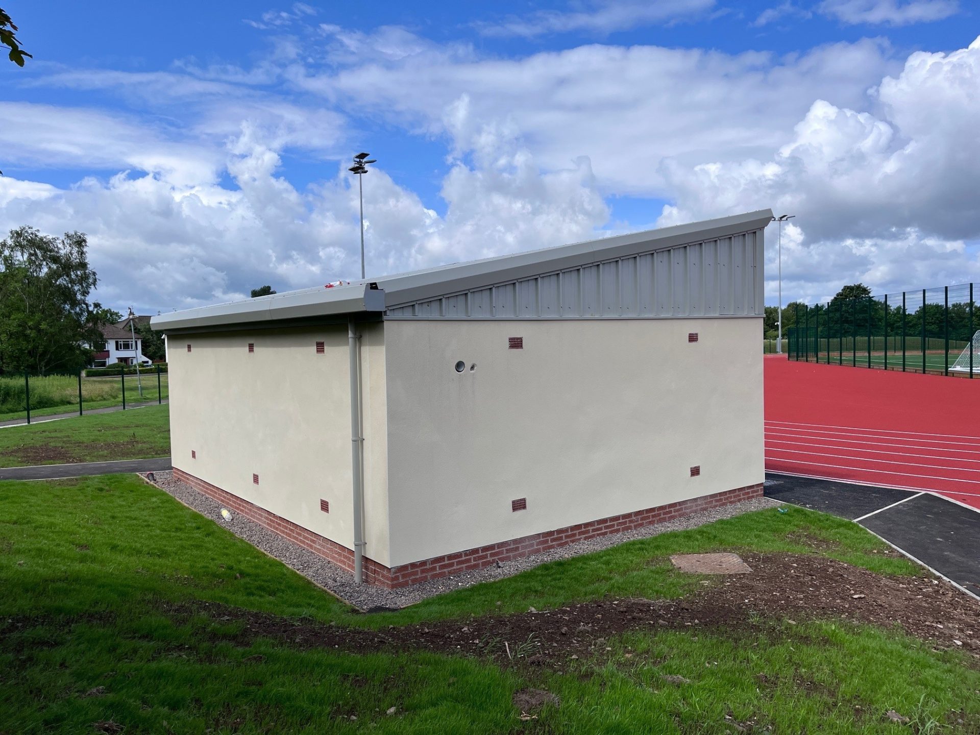 A modern sports facility features a sloped roof, light-colored walls, and a red athletic track and field visible in the background.