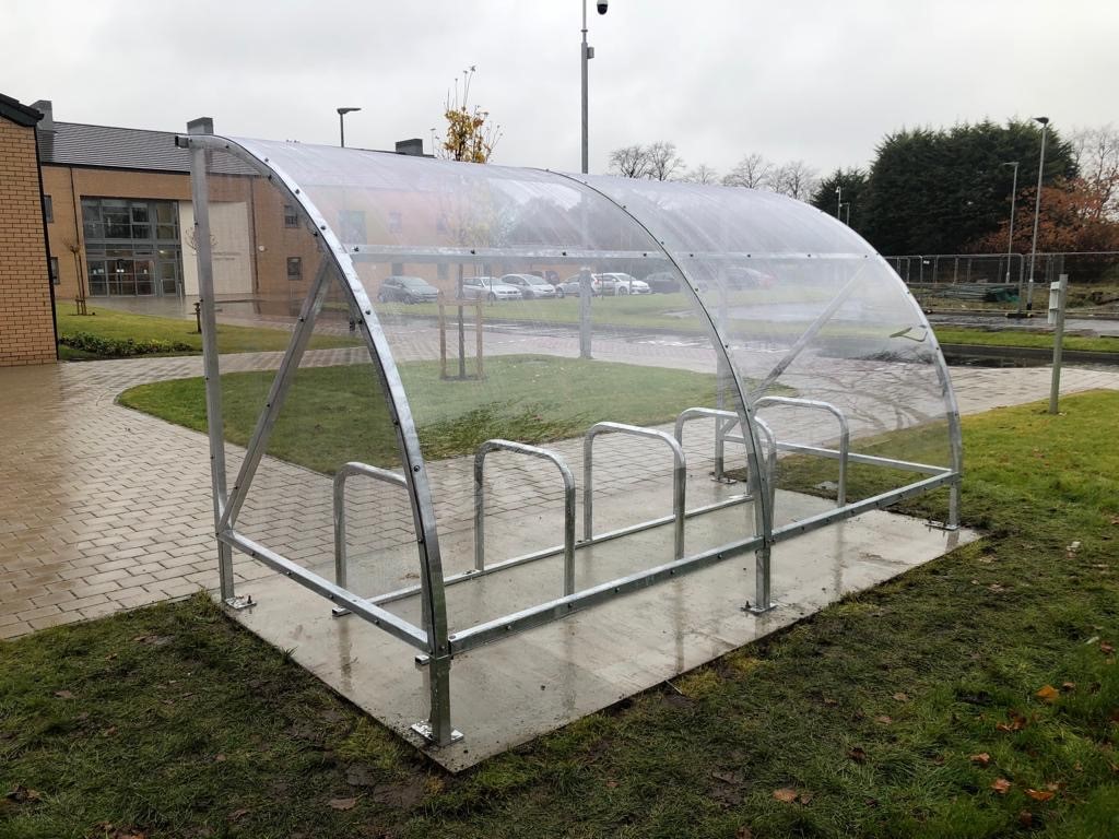 A transparent bicycle shelter with a metal frame, located on a paved area near a building, surrounded by grass and trees.