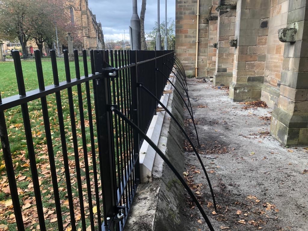 A black fence with angled supports lines a stone wall, surrounded by fallen leaves and green grass on a cloudy day.